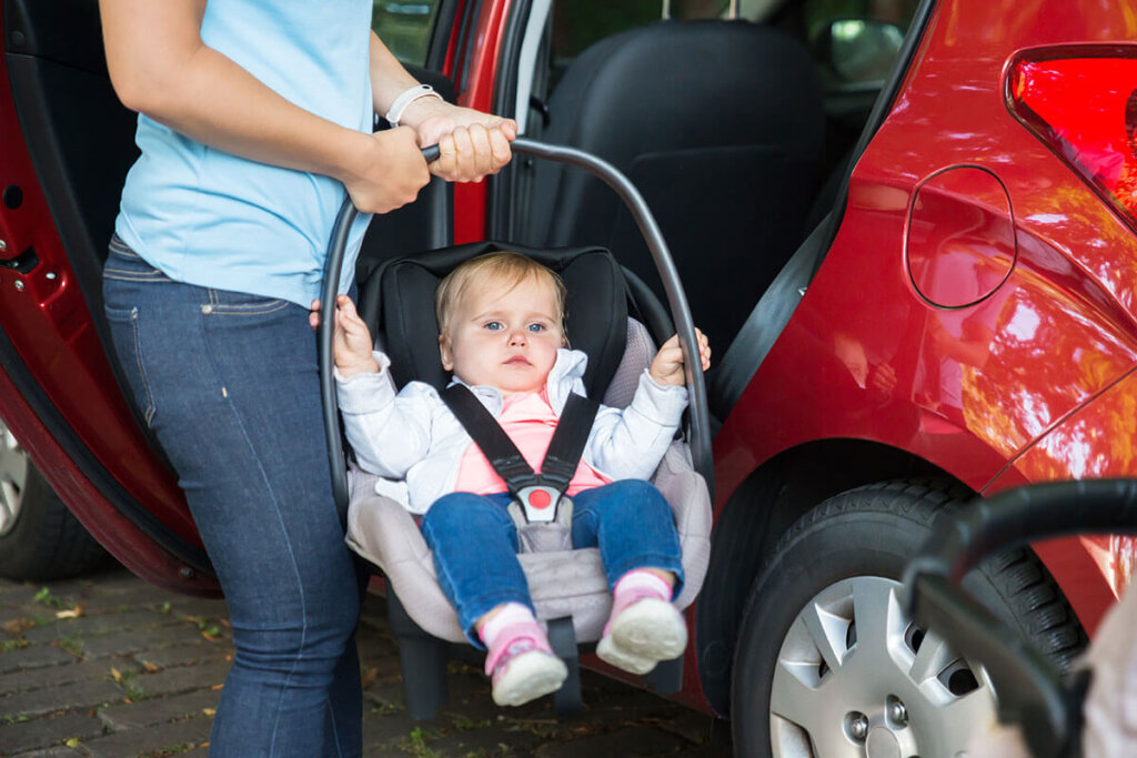 Une personne tient une coque bébé devant une voiture dont la portière est ouverte.